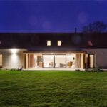 Osvetlená biela stavba s veľkou presklenou stenou a dreveným terasovým obkladom pod tmavou nočnou oblohou.