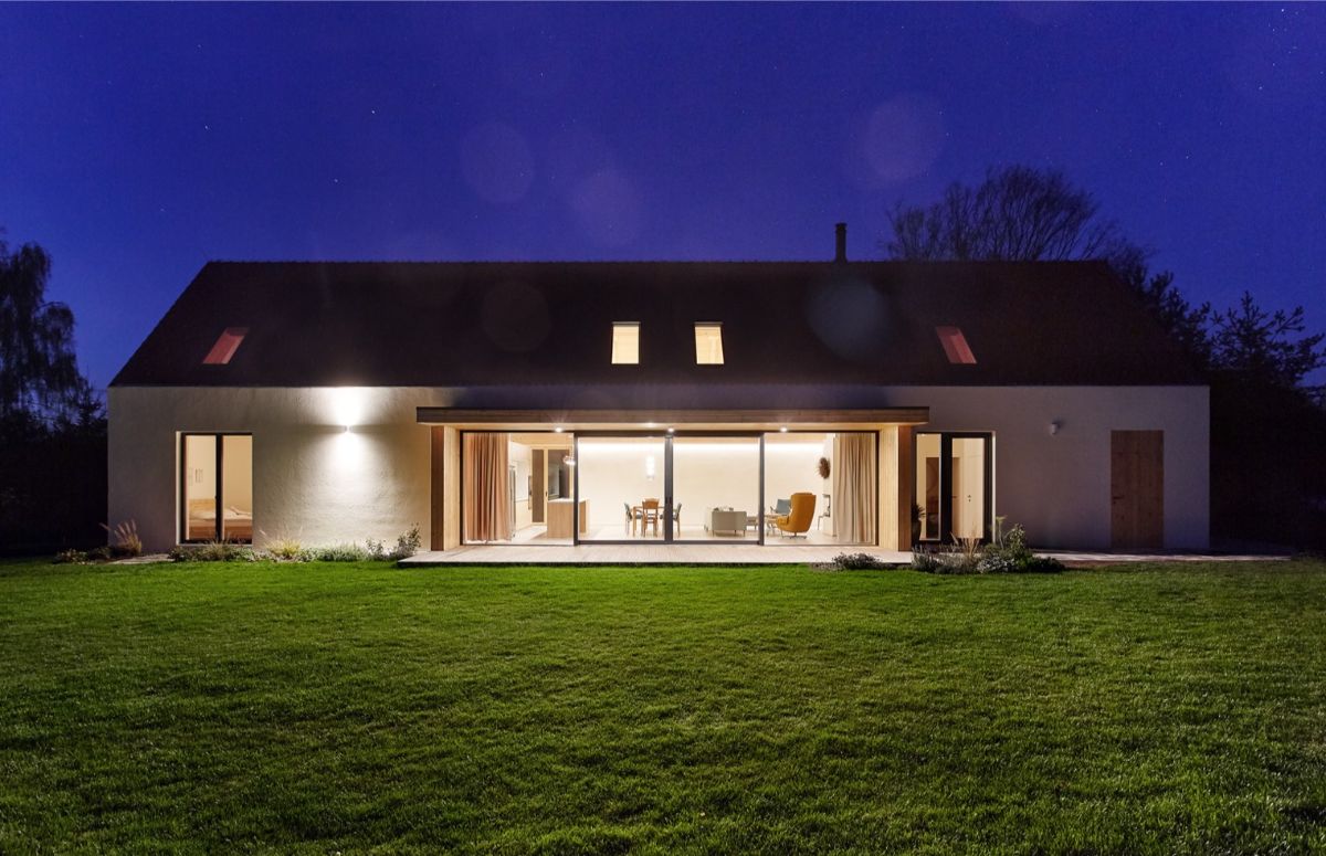 Osvetlená biela stavba s veľkou presklenou stenou a dreveným terasovým obkladom pod tmavou nočnou oblohou.