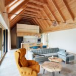 Loftový interiér s vysokým dreveným stropom, horčicovým ušiakom, dreveným konferenčným stolíkom a sivou dlažbou na podlahe.