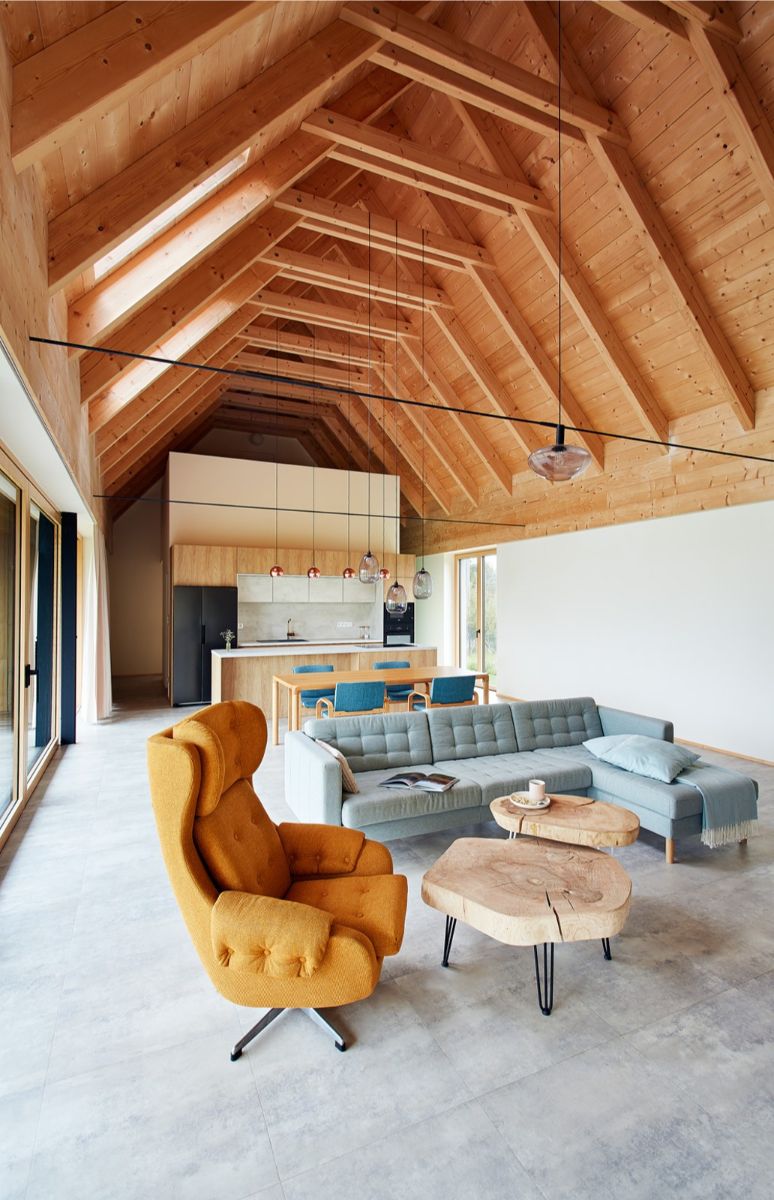 Loftový interiér s vysokým dreveným stropom, horčicovým ušiakom, dreveným konferenčným stolíkom a sivou dlažbou na podlahe.