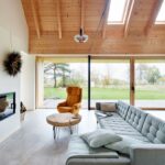 Obývacia izba s presklenou stenou, nízkou sivou pohovkou, dreveným stropom s priznanými trámami a bielym vstavaným kozubom.