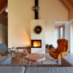 Minimalistický biely kozub s čiernou vložkou uprostred miestnosti, drevený konferenčný stolík a štýlové horčicové kreslo.