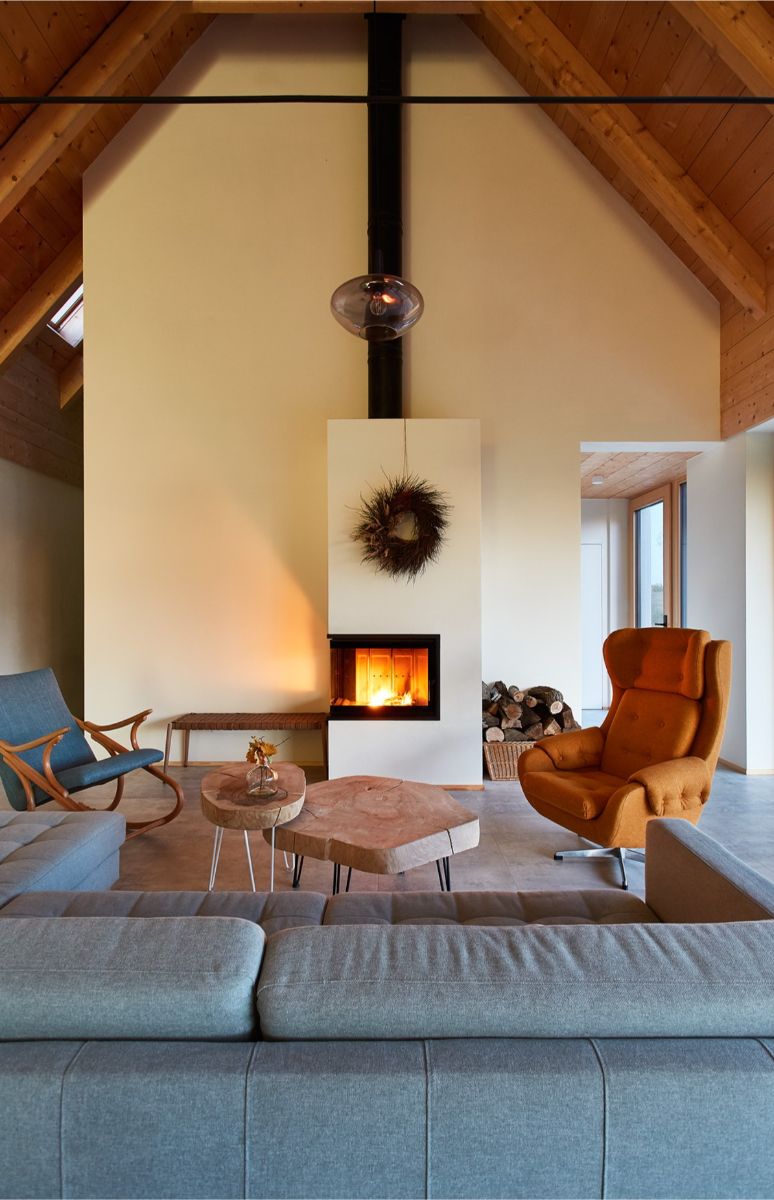 Minimalistický biely kozub s čiernou vložkou uprostred miestnosti, drevený konferenčný stolík a štýlové horčicové kreslo.