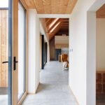 Svetlá chodba s betónovou dlažbou, bielymi stenami, dreveným podhľadom a výhľadom na presklené vchodové dvere do domu.