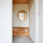 Úzka predsieň s drevenými policami z debničiek, okrúhlym zrkadlom s tenkým rámom a svetlou kamennou podlahou.