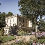 Historický zámok s rovnou strechou, obohnanou zábradlím v udržiavanej záhrade so stromami a kvetmi.