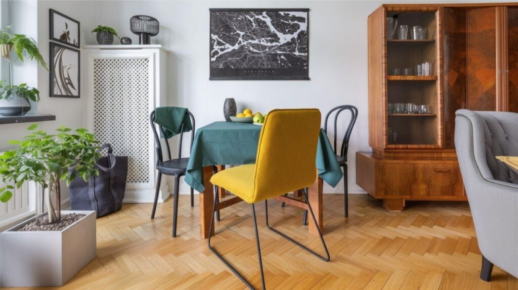 Žltá čalúnená stolička a dve zelené tonetky pri drevenom jedálenskom stole s tmavozeleným obrusom pred retro príborníkom.