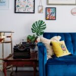 Čierny písací stroj na starožitnom drevenom stolíku vedľa kráľovsky modrej sedačky pod galériou obrazov.