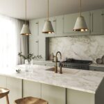 Biely mramorový ostrov s mosadznou batériou pred svetlozelenou kuchynskou linkou pod kamennými svietidlami.