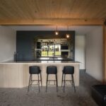 Moderný domáci bar s ryhovaným dreveným pultom, čiernymi koženými stoličkami a tmavosivou nikou s podsvietenými policami.