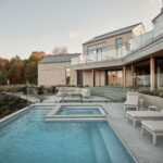 Príjazdová cesta k modernému domu so sivým šindľom, tmavou garážou, kovovou strechou a nízkym kamenným plotom.