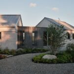 Vidiecky dom so svetlou fasádou, tmavým lamelovým prepojením, plechovou strechou a štrkovým chodníkom s brezami.