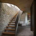 Interiérové schodisko s drevenými stupňami, sivým kovovým zábradlím, kamennou stenou a klenutým dreveným stropom.