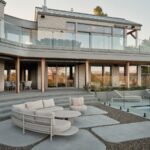 Terasa s krémovou oblou sedačkou, betónovou dlažbou, skleneným zábradlím a kamenným obkladom modernej vily.