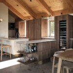 Kuchynská linka s tmavým dreveným dekorom, integrovanou vinotékou, kovovým barovým vozíkom a kamennou podlahou.