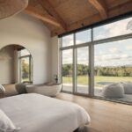 Svetlá spálňa s voľne stojacou kamennou vaňou, dreveným trámovým stropom a veľkoformátovým presklením do prírody.