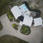 Letecký pohľad na moderný dom s tromi prepojenými krídlami, šedou plechovou strechou a dvorom s bazénom a ohniskom.