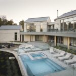 Exteriérová terasa so svetlou kamennou fasádou, modrým bazénom, bielymi ležadlami a preskleným balkónom s kovovou konštrukciou.