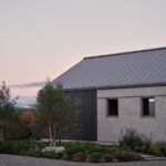 Detail fasády so sivým šindľovým obkladom, tmavým vertikálnym plechom, plechovou strechou a okrasnou výsadbou.