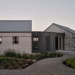 Vstupná časť domu s oblou stenou z tmavých lamiel, sivým obkladom a štrkovým chodníkom v zelenej záhrade.