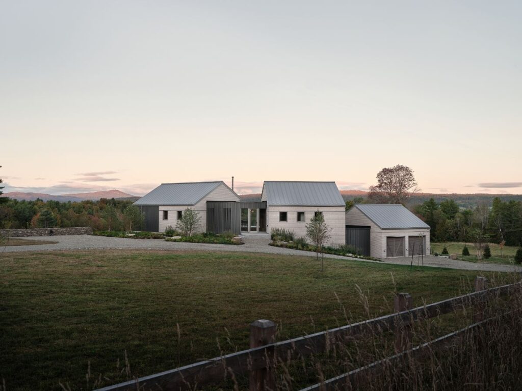 Príjazdová cesta k modernému domu so sivým šindľom, tmavou garážou, kovovou strechou a nízkym kamenným plotom.