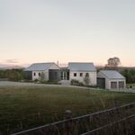 Príjazdová cesta k modernému domu so sivým šindľom, tmavou garážou, kovovou strechou a nízkym kamenným plotom.