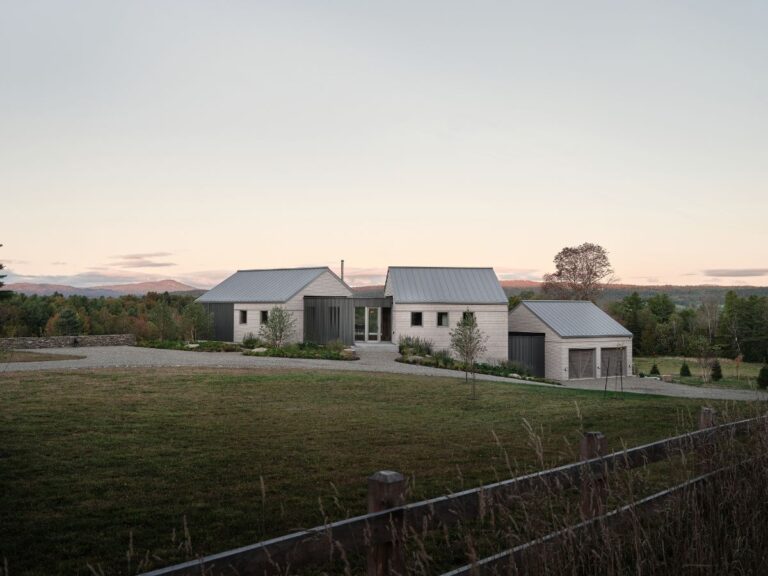 Príjazdová cesta k modernému domu so sivým šindľom, tmavou garážou, kovovou strechou a nízkym kamenným plotom.