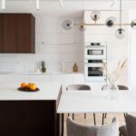 Moderná biela kuchyňa s tmavohnedou skrinkou, vertikálnym obkladom, ostrovom a mosadzným lustrom v minimalistickom štýle.