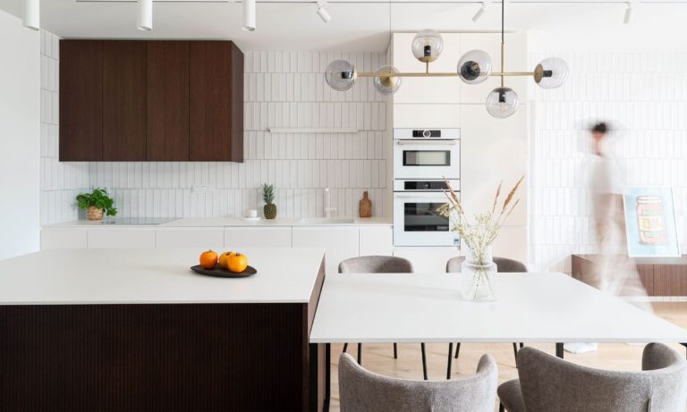 Moderná biela kuchyňa s tmavohnedou skrinkou, vertikálnym obkladom, ostrovom a mosadzným lustrom v minimalistickom štýle.