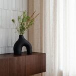 Čierna organická váza na tmavohnedej drážkovanej skrinke pri bielom obklade a svetlom ľanovom závese.