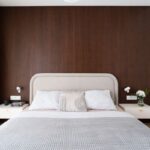 Posteľ s béžovým čalúneným čelom a biele samonosné nočné stolíky na tmavej lamelovej stene.