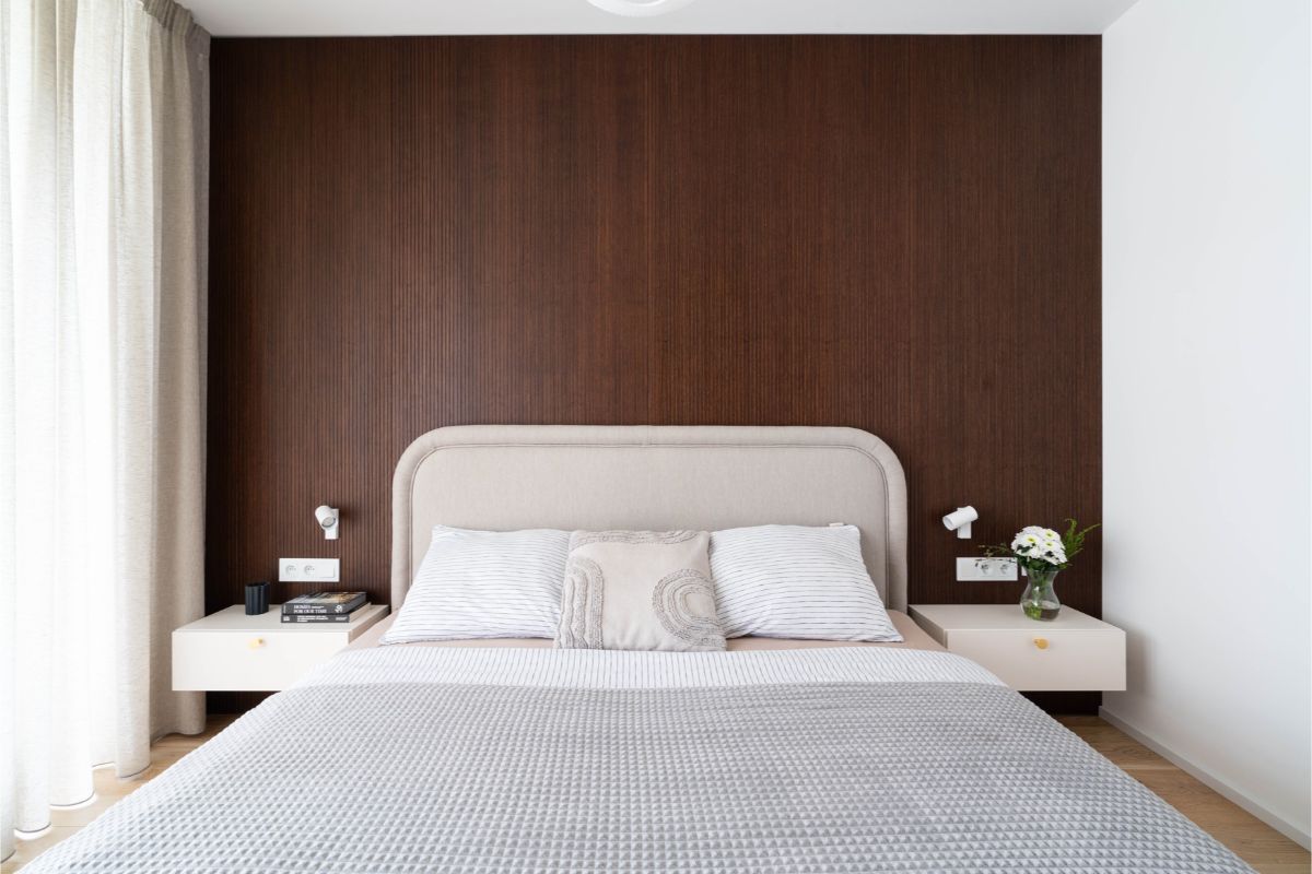 Posteľ s béžovým čalúneným čelom a biele samonosné nočné stolíky na tmavej lamelovej stene.