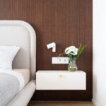 Biely samonosný stolík so zlatým úchytom na tmavom drevenom obklade pod minimalistickou lampou.