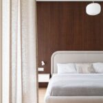 Svetlosivá posteľ pred tmavou dyhovanou stenou za béžovým ľanovým závesom.