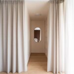 Oválne zrkadlo v svetlej šatníkovej chodbe lemovanej béžovými závesmi a dubovou podlahou.