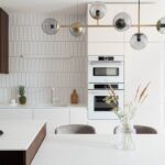 Biela kuchynská linka s tmavým dreveným akcentom, vstavanými rúrami a dizajnovým skleneným svietidlom nad jedálenským stolom.