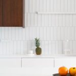 Biely vertikálny keramický obklad s bielym drezom, svetlou pracovnou doskou a lineárnym nástenným svietidlom.