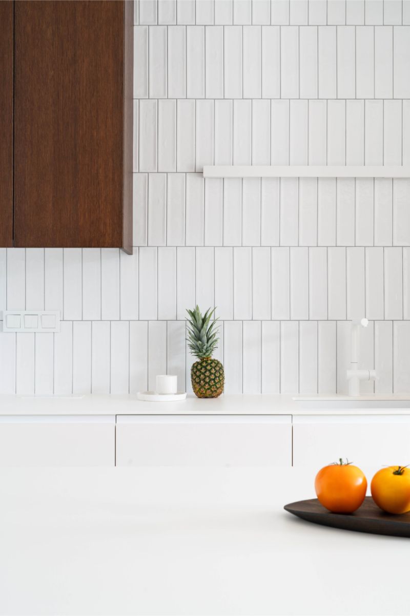 Biely vertikálny keramický obklad s bielym drezom, svetlou pracovnou doskou a lineárnym nástenným svietidlom.