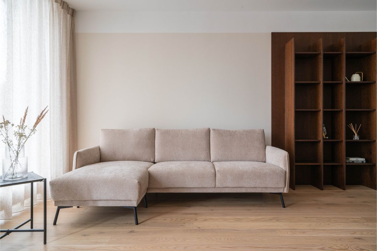 Béžová rohová sedačka na dubových parketách vedľa tmavohnedej vstavanej knižnice v minimalistickej obývačke.
