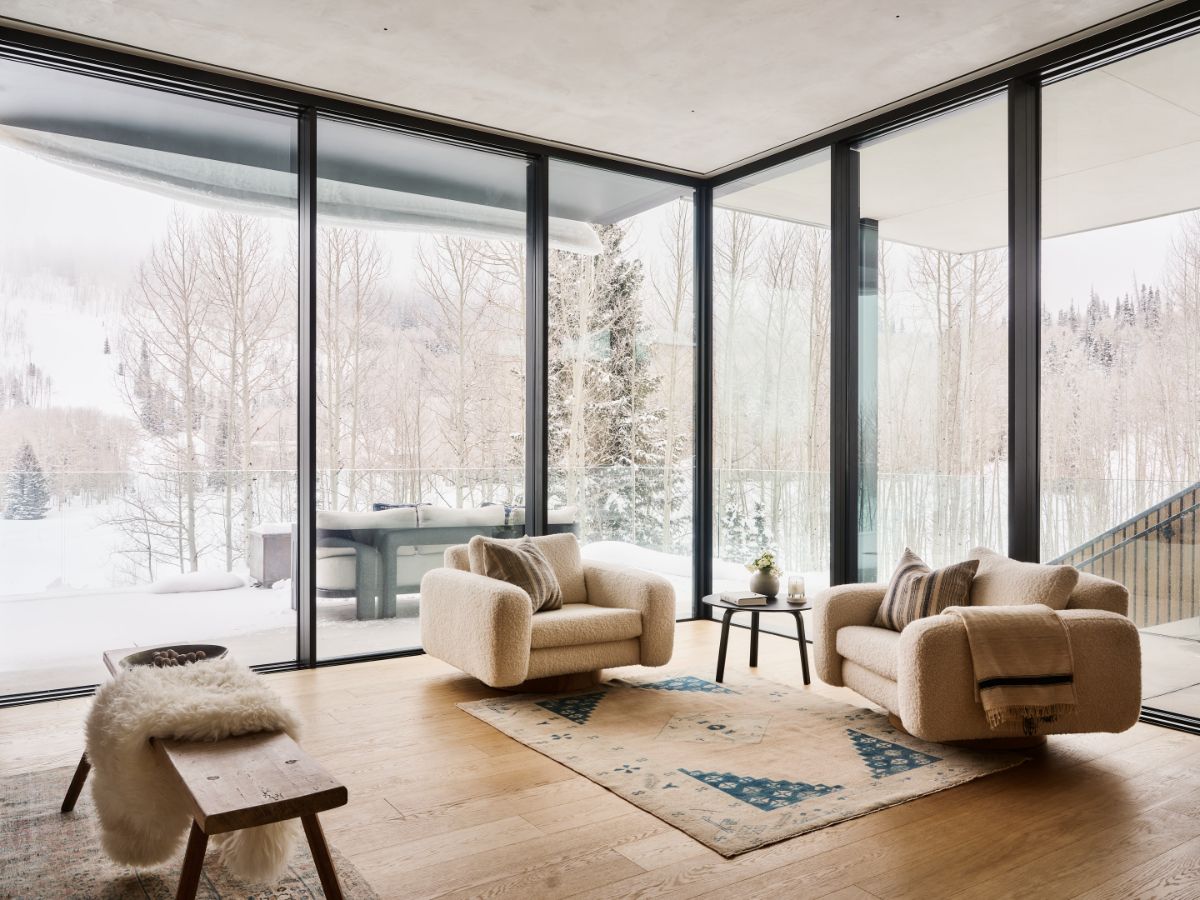 Svetlé krémové kreslá, drevená podlaha a veľkoformátové okná s výhľadom na zasnežené stromy v modernom interiéri.