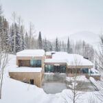 Exteriér modernej drevenej stavby s plochou strechou a presklenými fasádami v hlbokom snehu medzi ihličnatými stromami.
