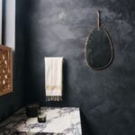 Mramorová umývadlová doska, tmavá štruktúrovaná stena a organické bezrámové zrkadlo v minimalistickej kúpeľni.