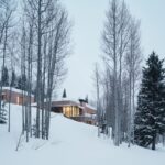 Horské sídlo s osvetlenými oknami, dreveným obkladom a zasnežená stráň s vysokými štíhlymi stromami za súmraku.