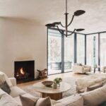 Otvorená obývacia izba s béžovou sedačkou, čiernym minimalistickým krbom a kovovým dizajnovým lustrom na strope.