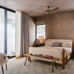 Moderná spálňa s čalúnenou posteľou, drevenou lavicou, hnedou omietkou a veľkými oknami s výhľadom na sneh.