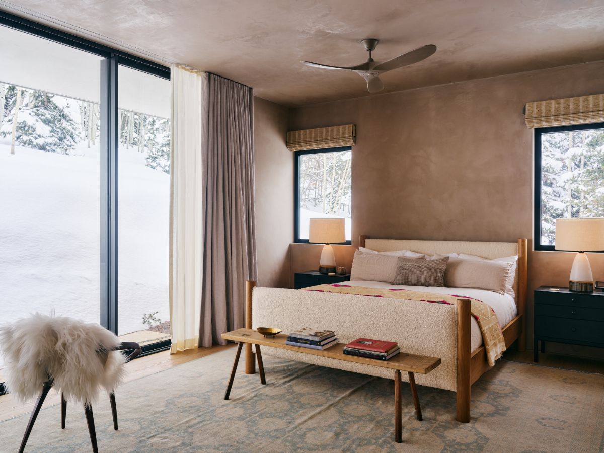 Moderná spálňa s čalúnenou posteľou, drevenou lavicou, hnedou omietkou a veľkými oknami s výhľadom na sneh.