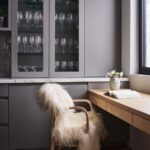 Sivý pracovný kút s dreveným stolom, kožušinovou stoličkou a presklenými skrinkami s čírymi pohármi na víno.