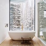 Samostatne stojaca biela vaňa pred veľkým oknom, drevený stolček a vzorovaný koberec v modernej kúpeľni.