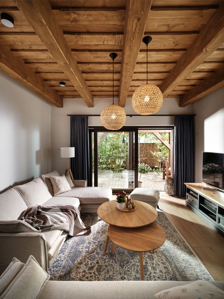 Svetlá obývacia izba s dreveným trámovým stropom, béžovou rohovou sedačkou a dvoma prútenými lustrami.