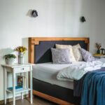 Manželská posteľ s masívnym dreveným čelom, biely nočný stolík a jemné vzorované vankúše vo svetlej spálni.